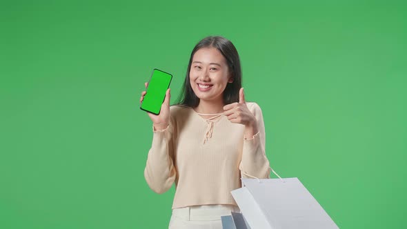 Asian Shopping Woman With Shopping Bags Holding Green Screen Mobile Phone Thumbs Up On Green Screen