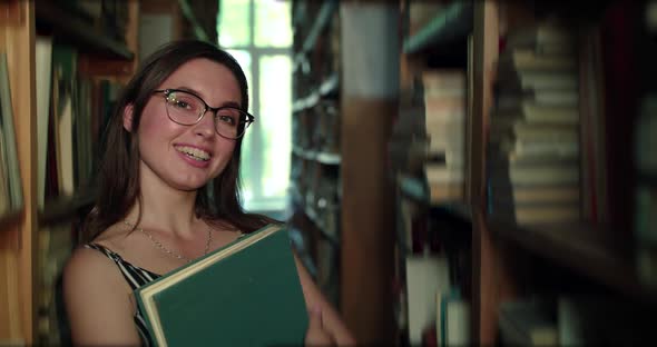 Girl in Glasses with Book in Hands Looking and Smiling at Camera in Library