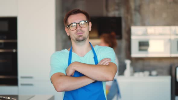 Portrait of Young Man Janitor in Apron Smiling at Camera with Hands Crossed Standing in Kitchen