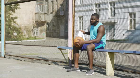 Sad Afro-American male sitting on the bench and playing ball, loneliness