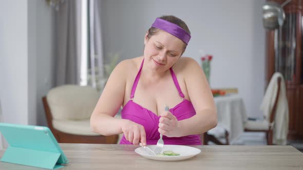 Motivated Obese Woman Cutting Cucumber Slices with Knife and Eating Healthful Veggie Food with Fork