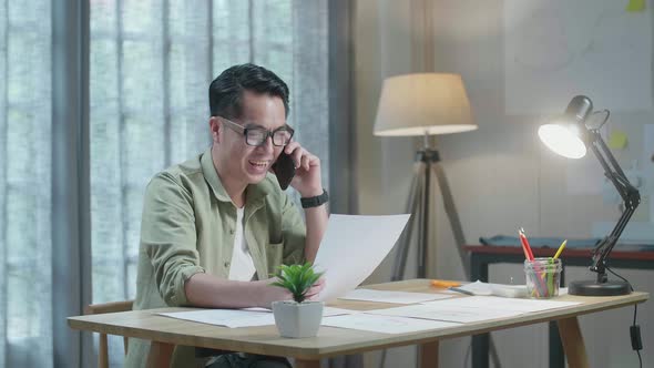 Asian Man Designer Talking On Smartphone While Looking At The Layout Bond In Hand At The Office