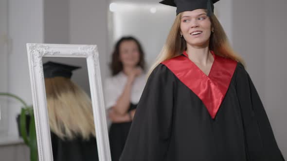 Happy Confident Young Woman in Graduation Gown and Cap Spinning Admiring Reflection in Mirror with