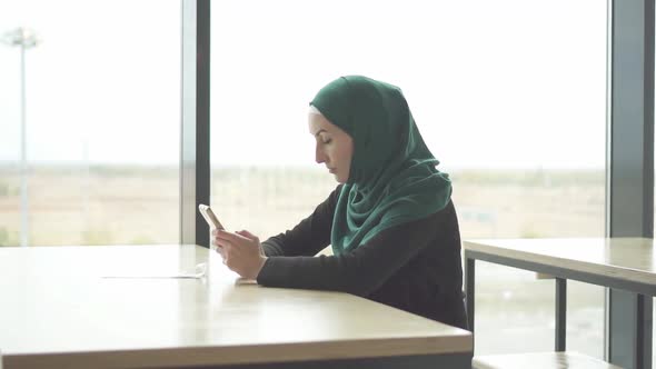Pretty Muslim Woman in Hijab Sitting at the Table and Uses the Phoneslow Mo