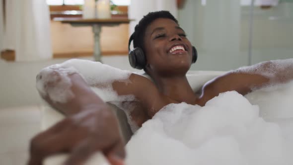 Smiling african american attractive woman wearing headphones relaxing in foam bath in bathroom