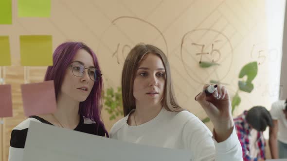 Caucasian Women Draw with a Marker on Glass Wall Looking at a Drawing Teamwork