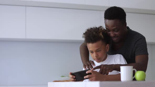 Young Father and Son are Using Smartphone While Sitting at Table in Home Kitchen Spbi