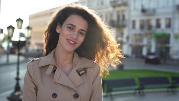 Portrait of Sunlit Curly Woman Wearing Coat Smiling While Strolling Through Morning City Street in