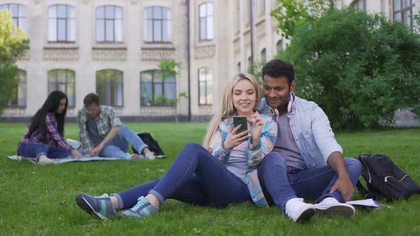 Loving Couple of Students Sitting on Lawn and Watching Video on Smartphone