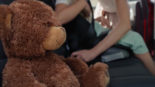 Teddy bear in the foreground and mother securing daughter into baby car seat in the background. Shot