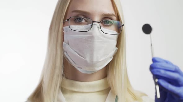 Young Female Dentist in Mask Approaches With Tools Holding a Mouth Mirror and Dentist's Hook