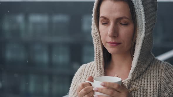 Caucasian Woman Stays on Balcony During Snowfall with Cup of Hot Coffee or Tea