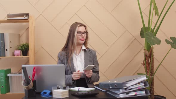 Woman Office Worker Sits at Desk with Laptop and Several Large Folders of Papers and Shakes Head in