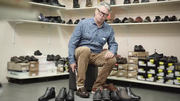 Stubborn Senior Man Trying on Too Small Boot in Shoe Store. Wide Shot of Elegant Caucasian Male