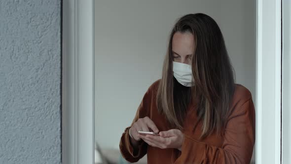 Woman in orange sweater and medical mask using phone by the window indoors