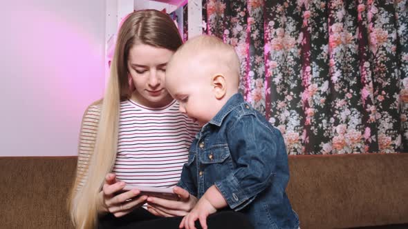 A Young Mother and Her Young Son Sit on the Couch and Look at Pictures on Their Phone