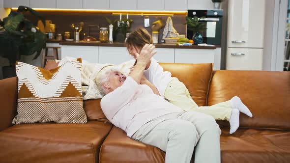 Grandma is Laughing Lying on Sofa and Playing with Her Little Granddaughter in Studio Apartment with