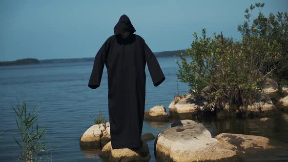Woman devil in black cape and hood on stones near the river. Halloween costume.