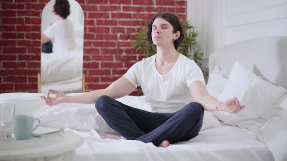 Wide Shot Portrait of Confident Calm Young Man Sitting on Bed in Lotus Pose and Closing Eyes