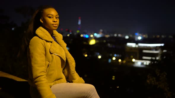 A Young Black Woman Looks Around and at the Camera As She Sits on a Low Wall in Urban Area at Night