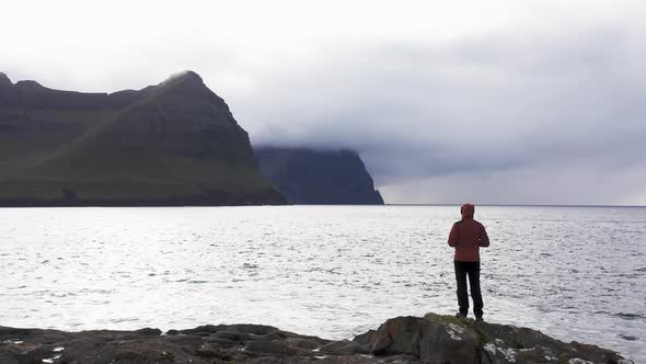 Aerial View of Unrecognizable Woman Stands on Rock Cliff Looking at Wilderness Background of Faroe