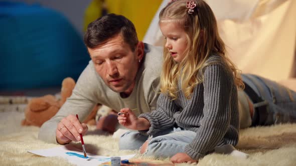 Cute Little Girl Painting with Man Lying at Tent in Living Room at Home