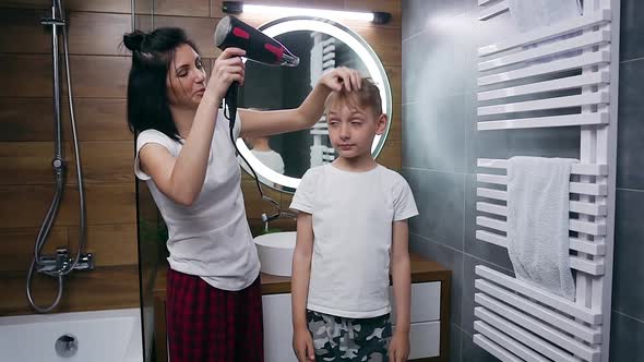 Boy Standing Near the Bathroom Mirror While His Adorable Mother Drying His Hair with Hair Dryer