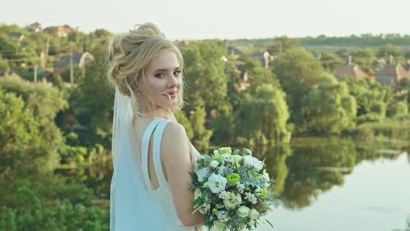 Happy Young Woman Bride with a Wedding Bouquet Stands on the Background of the River