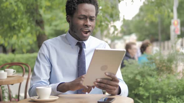 African Businessman Celebrating Win on Tablet Sitting in Outdoor Cafe