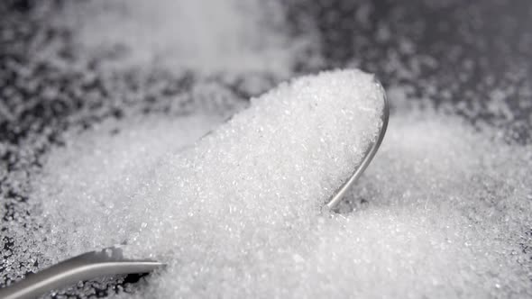 A full teaspoon with white sugar. Falling crystals into a spoon and over a kitchen slate surface