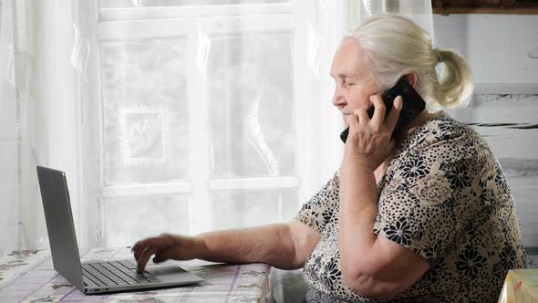 Beautiful Gray-haired Woman with Phone and Laptop Sits at a Table By the Window