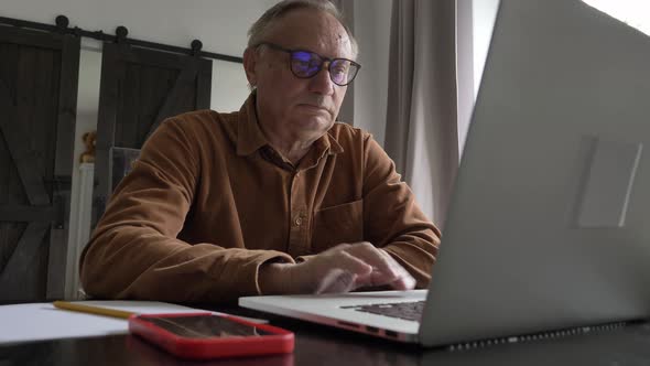 Old man in eyeglasses using laptop computer at home.