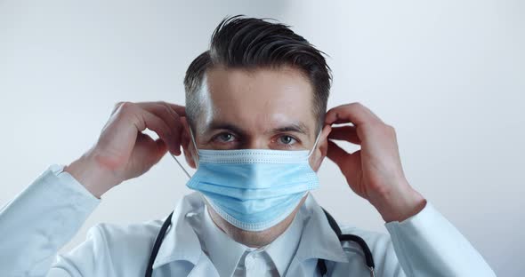 Portrait of Male Doctor Putting on Medical Mask and Looks at the Camera Over White Background