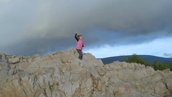 Cute girl in a pink vest stands on a cliff above the sea at sunset