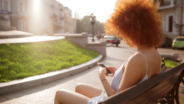 Young Beautiful Girl Typing on Phone Sitting on Bench in City Park