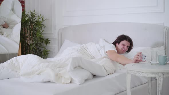 Wide Shot of Sad Dissatisfied Man Turning Off Ringing Alarm Clock and Closing Head with Pillow