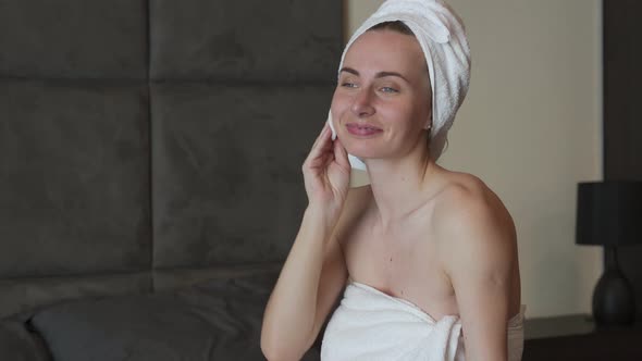 Young Brunette with a Towel on Her Head Removes Makeup From Her Face While Sitting on the Bed