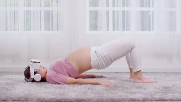 Happy Asian Pregnant woman using virtual reality glasses for practice yoga online class Half bridge