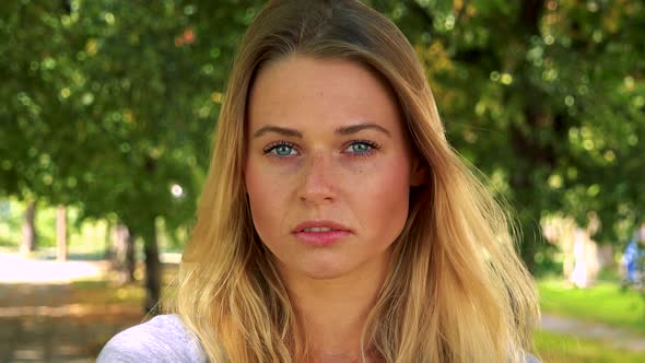 Young Pretty Blond Woman Looks To Camera with Serious Face - Park with Trees in Background - Closeup