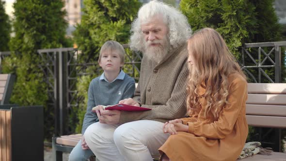 Angle View of Intelligent Male Retiree Explaining Book To Grandchildren on Sunny Day Outdoors