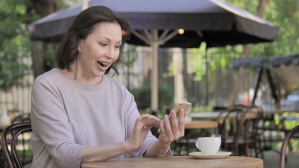 Old Woman Cheering Success on Smartphone Sitting in Outdoor Cafe