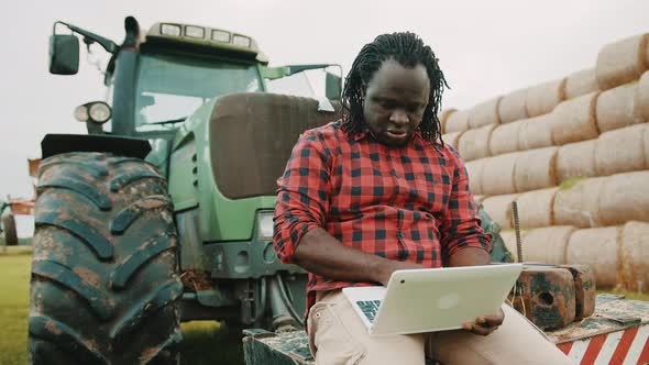 Young African Farmer Using Tablet While Sitting on Green Tractor. Haystack in the Background