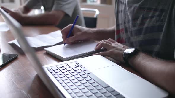 Close-up of man writing in notebook with laptop computer nearby