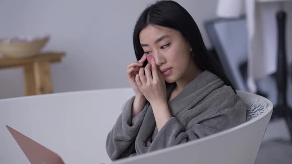 Confident Beautiful Asian Young Woman Applying Eye Patches Sitting in Bathtub with Laptop