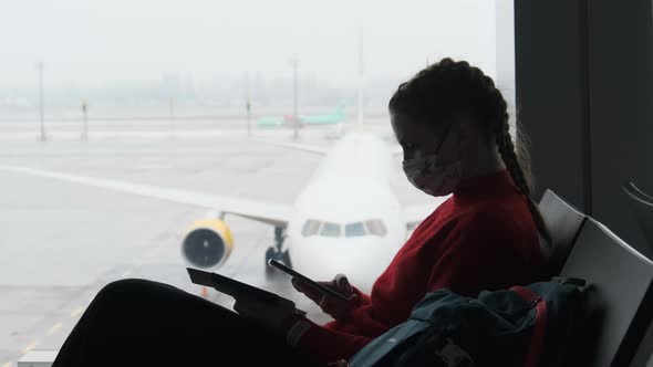 Masked Young Woman in Airport Waiting Room Sits and Using a Smartphone By Window