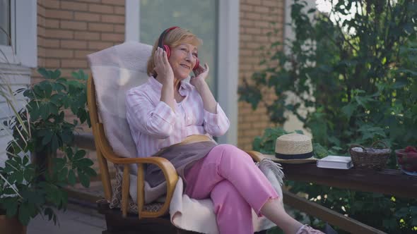 Smiling Charming Senior Caucasian Woman Listening to Music in Headphones Sitting on Porch on Front