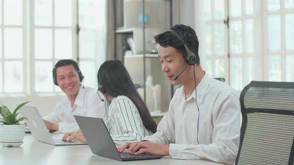 A Man Of Three Asian Call Centre Speaking To Customers While His Colleagues Talking With Each Other
