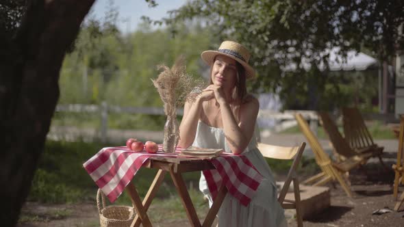 Young Girl in a Straw Hat and White Dress Sitting at the Small Table and Looking at the Camera
