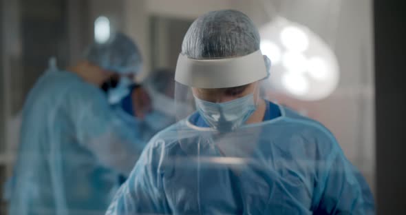 Portrait of Male Surgeon Standing Near Glass Door While Team Performing Surgery in Operation Room
