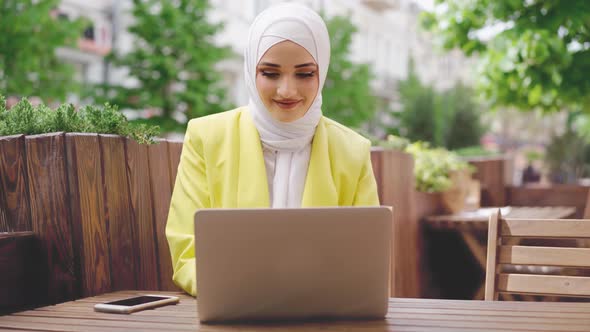 Smiling Young Muslim Woman Wearing Headscarf Sits in Cafe and Uses Laptop